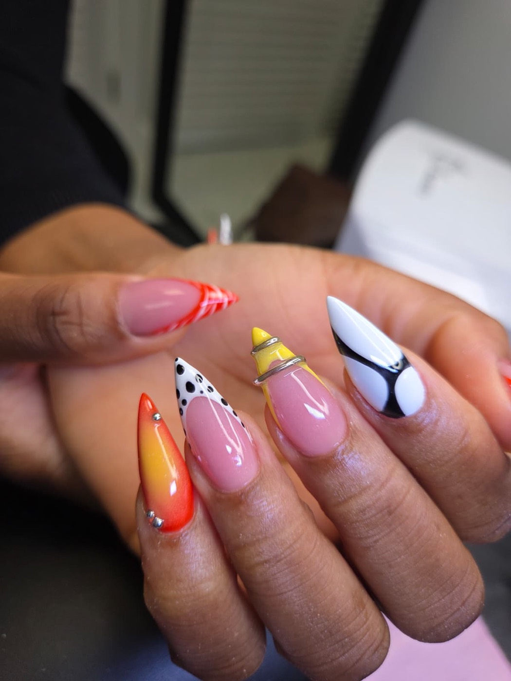 Almond nails with mixed designs — orange and yellow ombré, black polka-dot French, and a black and white swirl